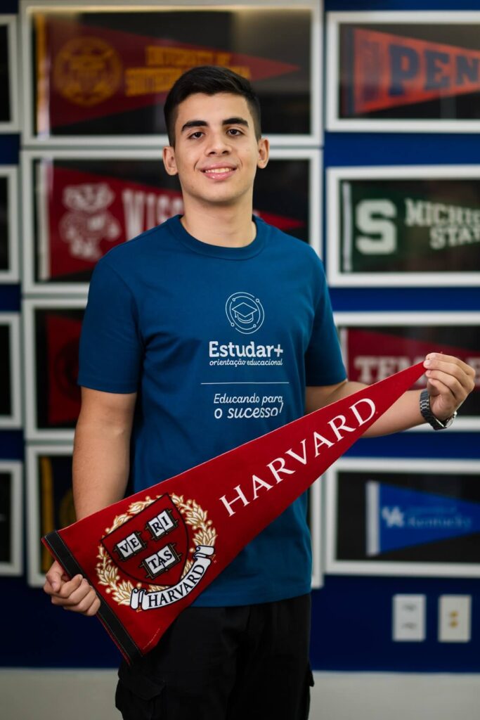 Estudante com camiseta da Estudar+ segurando flâmula oficial de aprovação na Universidade de Harvard